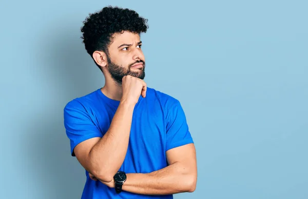 あごの質問について考える上で手でカジュアルな青のTシャツを着てひげを持つ若いアラブ人男性は 集中的な表現 思慮深い顔で笑顔 疑わしい概念 — ストック写真