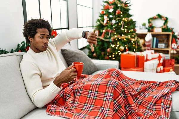 Young african american man sitting on the sofa drinking coffee by christmas tree looking unhappy and angry showing rejection and negative with thumbs down gesture. bad expression.