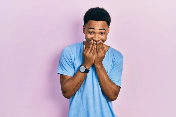 若いアフリカ系アメリカ人男性を着てカジュアルブルーTシャツ笑いと恥ずかしがり屋のクスクス笑いをカバー口で手 ゴシップやスキャンダルの概念 — ストック写真