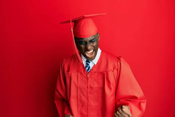卒業キャップと儀式用のローブを身に着けている若いアフリカ系アメリカ人男性は 腕を上げて 笑顔と成功のために叫んで勝者のジェスチャーを行うことは非常に幸せで興奮しています お祝いのコンセプト — ストック写真