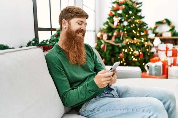 Ung Rödhårig Man Med Smartphone Sitter Vid Julgranen Hemma — Stockfoto