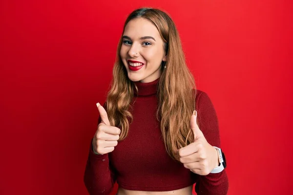 若いブロンドの女性は 手で積極的なジェスチャーを行うタートルネックセーターの成功サインを身に着けています 親指アップ笑顔と幸せ 陽気な表情と勝者のジェスチャー — ストック写真
