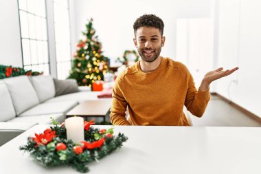 Arap genç adam, Noel ağacının yanında oturmuş gülümsüyor, avucunun içinde kameraya bakıyor ve gülümsüyor.. 