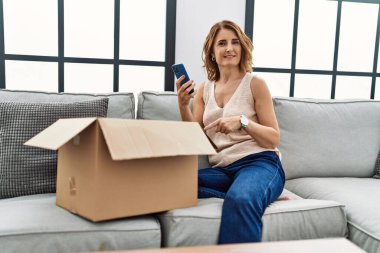 Orta yaşlı bir kadın kanepede oturmuş, teslimat paketini kontrol ediyor ve gülümsüyor. 