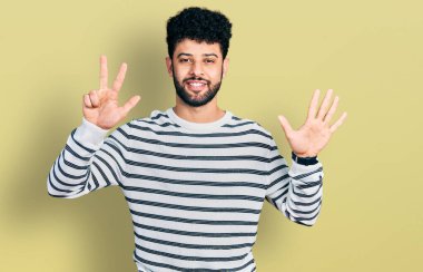 Sakallı, sıradan çizgili süveter giyen genç bir Arap sekiz numaralı parmaklarıyla gülümseyerek kendinden emin ve mutlu bir şekilde onu işaret ediyor.. 