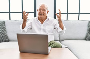 Evde dizüstü bilgisayar kullanan yaşlı adam koltukta çok mutlu ve heyecanlı bir şekilde oturuyor. Kolları havada, gülümseyerek ve başarı için çığlık atarak kazanan jestler yapıyor. kutlama konsepti. 