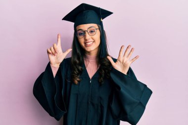 Mezuniyet şapkası ve tören cübbesi takan genç İspanyol kadın yedinci parmaklarını gösterip gösterirken kendinden emin ve mutlu bir şekilde gülümsüyor.. 
