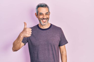 Yakışıklı, orta yaşlı, gri saçlı, sıradan tişörtlü, mutlu ve pozitif gülümseyen, başparmağıyla mükemmel ve onay işareti yapan bir adam. 