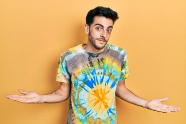 Sıradan kıyafetler giyen, elleri ve kolları havada, kafası karışmış genç İspanyol bir adam. şüphe kavramı. 