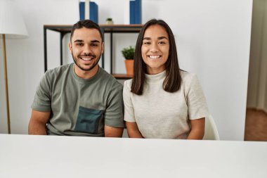 Genç Latin çift, gündelik giysiler içinde masada oturmuş mutlu ve pozitif görünüyor. Dişlerini gösteren kendine güvenen bir gülümsemeyle duruyorlar. 