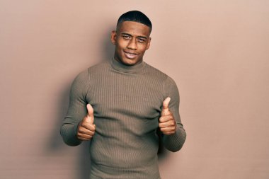 Balıkçı yaka kazak giyen genç bir zenci. Başarı işareti ile el hareketi yapıyor. Gülümsüyor ve mutlu. Neşeli ifade ve kazanan jest. 