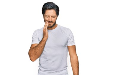 Günlük beyaz tişört giyen genç İspanyol adam diş ağrısı ya da diş ağrısı yüzünden acı dolu bir ifadeyle ağıza dokunuyor. dişçi 