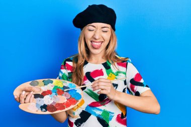 Kafkasyalı genç kadın sanatçı elinde fırça ve palet tutarak komik bir ifadeyle dilini dışarı çıkarıyor.. 