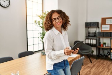 Orta yaşlı İspanyol kadın, ofiste dokunmatik ped kullanarak çalışırken kendine güvenerek gülümsüyor.