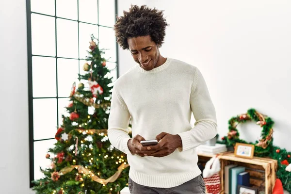 Young african american man uisng smartphone standing by christmas tree at home.