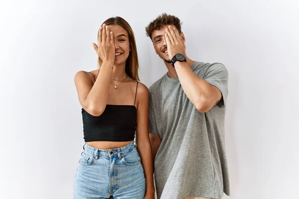 Young beautiful couple standing together over isolated background covering one eye with hand, confident smile on face and surprise emotion.