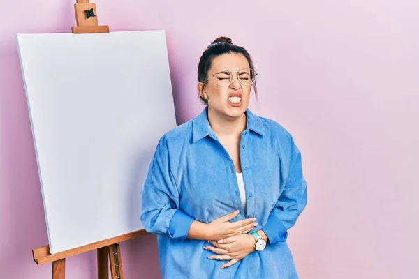 若いヒスパニック系の女性の画家のイーゼルで胃の上に手で立っているため 消化不良 痛みを伴う病気の気分が悪い キャッシュ コンセプト — ストック写真