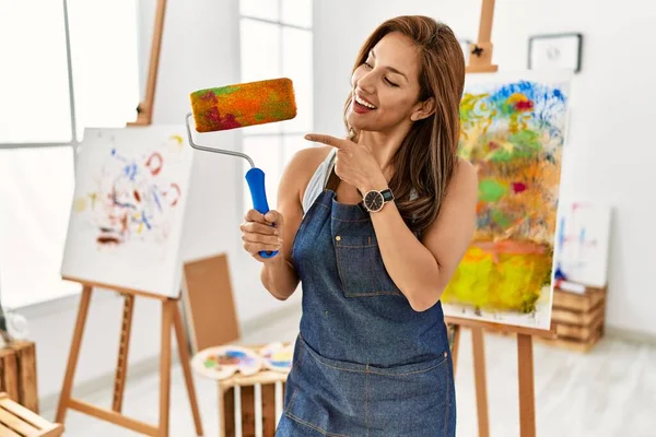 年轻的拉丁艺术家女人拿着颜料滚筒站在艺术工作室 高兴地微笑着 手指手画脚 — 图库照片