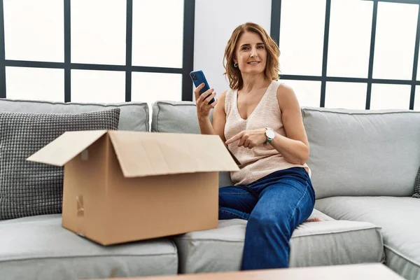 Orta yaşlı bir kadın kanepede oturmuş, teslimat paketini kontrol ediyor ve gülümsüyor. 