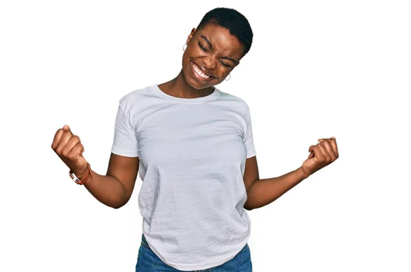 若いアフリカ系アメリカ人の女性は カジュアルな白いTシャツを着て非常に満足し 腕を上げ 笑顔と成功のために叫んで勝者のジェスチャーを行う興奮しています お祝いのコンセプト — ストック写真
