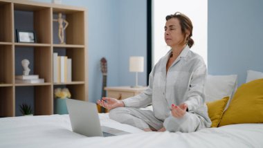 Orta yaşlı İspanyol kadın yatak odasında online yoga yapıyor.
