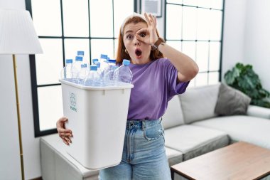 Elinde plastik şişelerle çöp sepetini geri dönüşüm sepetine koyan genç kızıl saçlı kadın şaşırmış yüz ve gözlerle parmaklarından bakıyor. İnançsız ifade. 