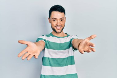 Gündelik giysiler giymiş genç İspanyol adam kameraya bakıp sarılmak için kollarını açmış gülümsüyor. Mutluluğu kucaklayan neşeli bir ifade.. 