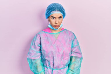 Genç esmer kadın cerrah üniforması giyiyor ve tıbbi maske takıyor şüpheci ve gergin, sorun yüzünden surat asıyor. negatif kişi. 