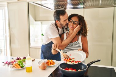 Orta yaşlı İspanyol çift, gülümsüyor. Mutfakta tavaya yiyecek koyuyorlar.