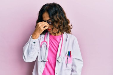 Orta yaşlı İspanyol kadın doktor üniforması giyiyor ve gözlük takıyor. Burnunu ve gözlerini ovuşturup yorgunluk ve baş ağrısı çekiyor. Stres ve hüsran kavramı. 