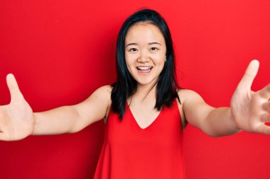 Sıradan kıyafetler giymiş genç Çinli bir kız kameraya bakıp sarılmak için kollarını açmış gülümsüyor. Mutluluğu kucaklayan neşeli bir ifade.. 