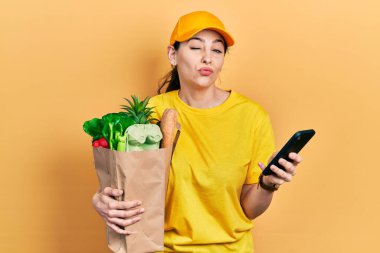 Genç İspanyol kadın elinde alışveriş poşeti tutarken akıllı telefon kullanıyor kameraya bakıp öpüşürken sevimli ve seksi oluyor. aşk ifadesi. 