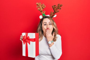 Orta yaşlı, beyaz, şirin Noel geyiği boynuzları takan, ağzı eliyle kapatan ve hata yapmaktan korkan bir kadın. şaşırmış ifade 