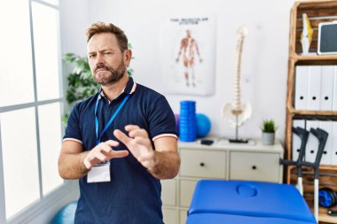 Orta yaşlı fizyoterapist. Ağrı giderme kliniğinde çalışıyor. Tiksinme reaksiyonu yüzünden tiksinmiş ve korkmuş. Eller havada. 