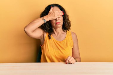 Orta yaşlı, sıradan kıyafetler giyen güzel bir kadın masaya oturmuş, gözlerini eliyle kapatıyor, ciddi ve üzgün görünüyor. Görme, saklama ve reddetme konsepti 