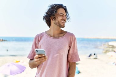 Genç İspanyol adam sahilde akıllı telefon kullanarak mutlu bir şekilde gülümsüyor..