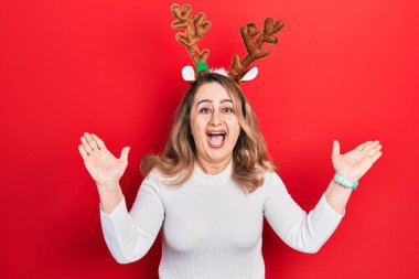 Orta yaşlı beyaz kadın şirin Noel geyiği boynuzları takıyor çılgınlığı kutluyor ve başarıya hayret ediyor kollar havada ve gözleri açık heyecanla haykırıyor. Kazanan konsept 