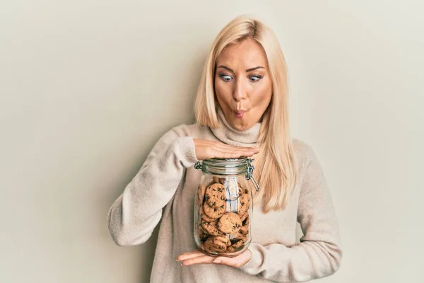Young blonde woman holding jar with chocolate chips cookies making fish face with mouth and squinting eyes, crazy and comical.