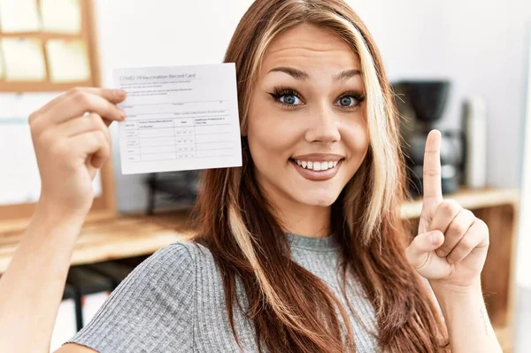 Young caucasian woman holding covid record card surprised with an idea or question pointing finger with happy face, number one