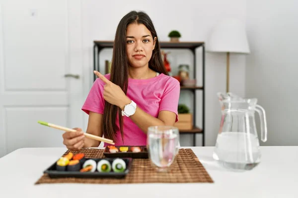 年轻的黑发女人用筷子吃寿司 用手指头指着旁边的广告 严肃而沉着 — 图库照片