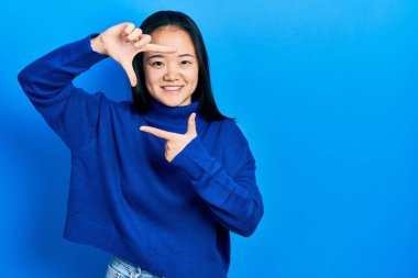 Gündelik kıyafetler giyen genç Çinli kız gülümseyerek elleri ve parmakları gülen yüzüyle çerçeve yapıyor. yaratıcılık ve fotoğrafçılık kavramı. 