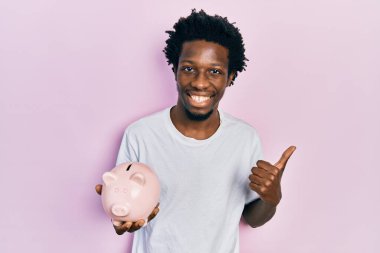 Genç Afrikalı Amerikalı domuz kumbarası tutan mutlu ve pozitif gülümseyen, başparmağıyla mükemmel ve onay işareti yapan adam. 