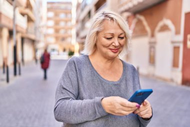 Orta yaşlı sarışın kadın sokakta akıllı telefon kullanarak gülümsüyor.