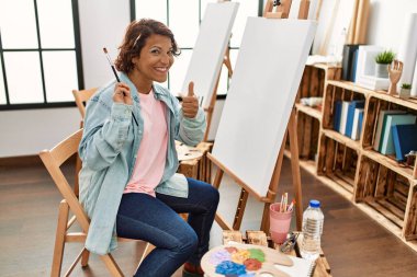 Orta yaşlı İspanyol bir kadın resim stüdyosunda tuvale resim yapıyor mutlu ve pozitif gülümsüyor, başparmağıyla mükemmel ve onay işareti yapıyor. 