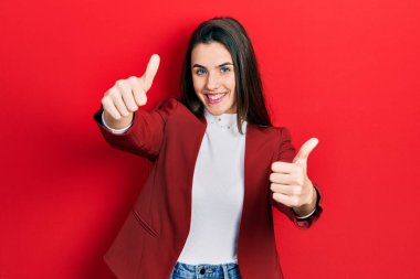 İş ceketi giyen genç esmer kız elleriyle olumlu jest yapmayı onaylıyor, gülümsüyor ve başarı için seviniyor. kazanan jest. 