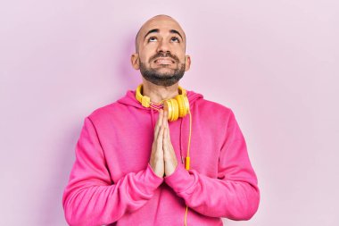 Spor kıyafetleri giyen ve kulaklık takan genç kel bir adam elleri bağlı yalvarıyor ve dua ediyor. Yüzünde umut ifadesi var. Çok duygusal ve endişeli. Yalvarıyor. 