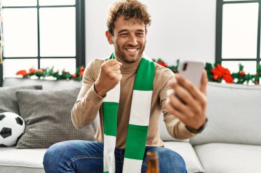 Genç İspanyol adam akıllı telefon kullanarak futbol maçını destekliyor evde Noel dekorunun yanında oturuyor.