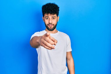 Sakallı, beyaz tişörtlü genç bir Arap, kameraya kızgın ve sinirli bir şekilde bakıyor. 