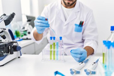 Laboratuvarda pipet ve test tüpü kullanarak bilim adamı üniforması giyen İspanyol genç bir adam.