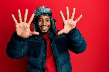 Sakallı, kışlık şapkalı ve paltolu genç Afrikalı Amerikalı on numaralı parmaklarıyla onu gösterirken kendinden emin ve mutlu gülümsüyor.. 